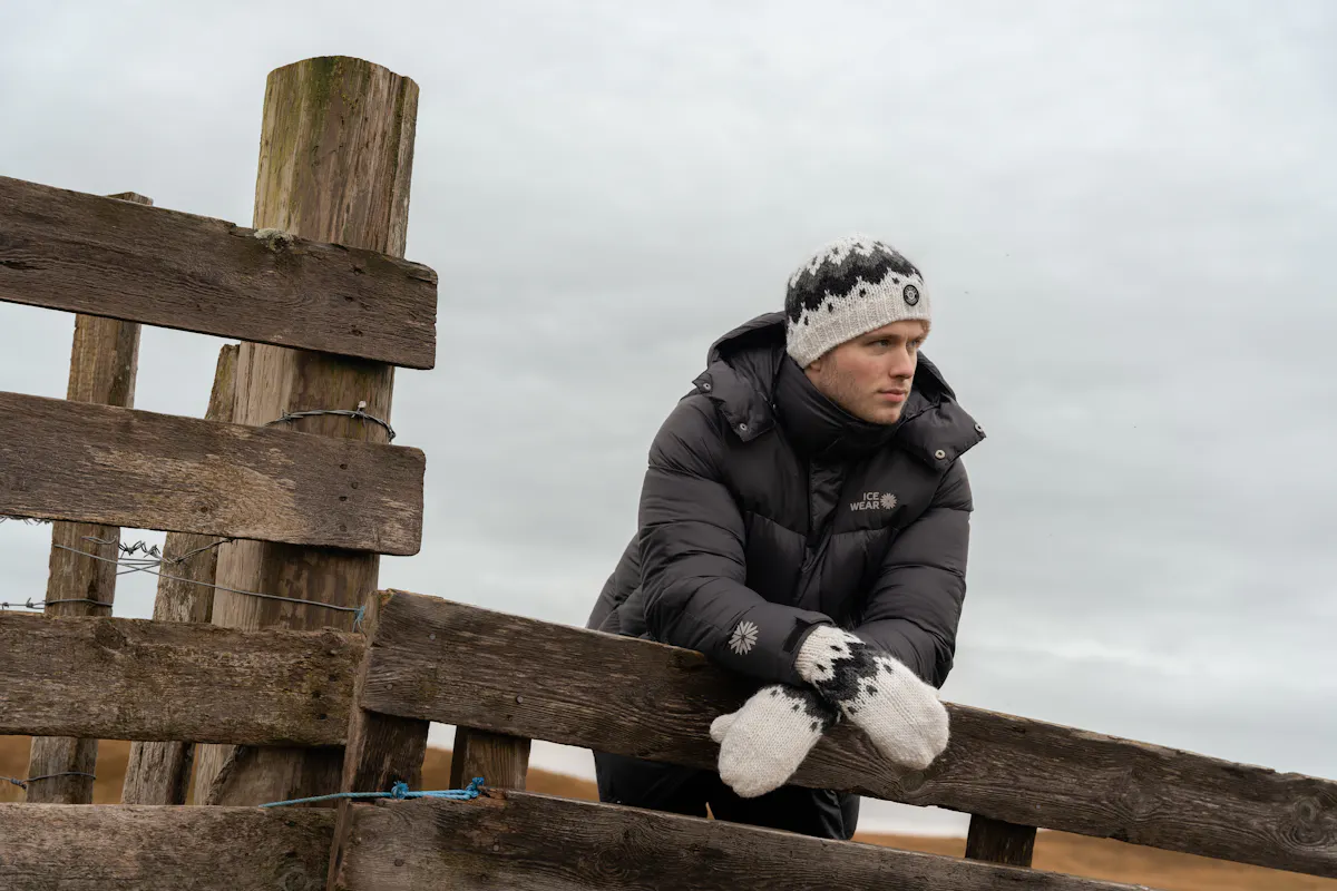 A man standing by a fence, wearing a warm coat with a hat and mittens