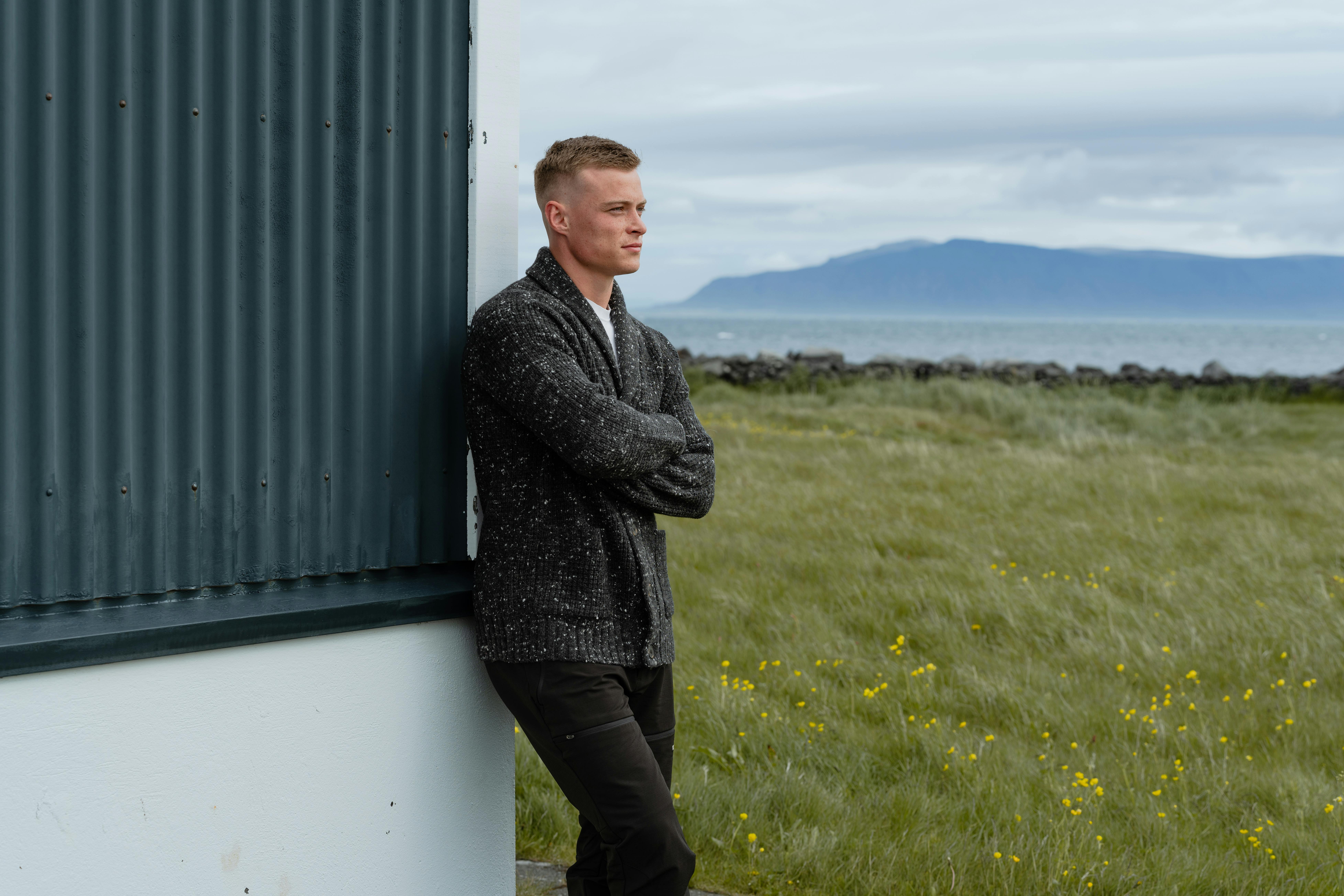 man standing next to old house, the sea and mountain behind him