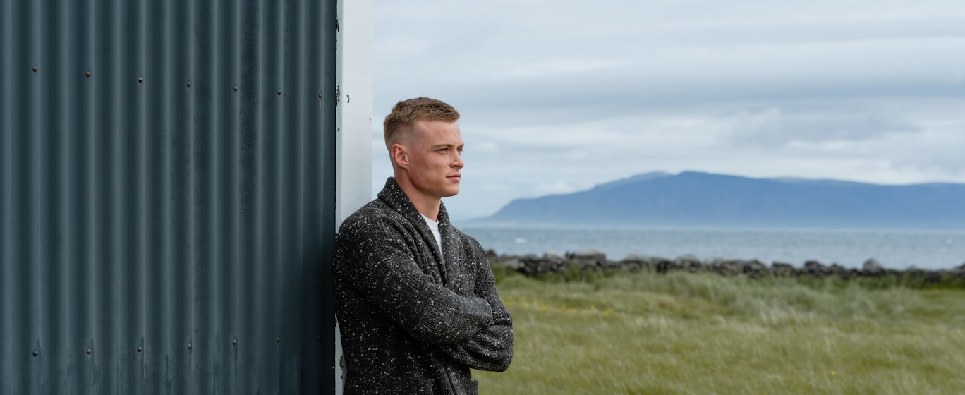 man standing next to old house, the sea and mountain behind him