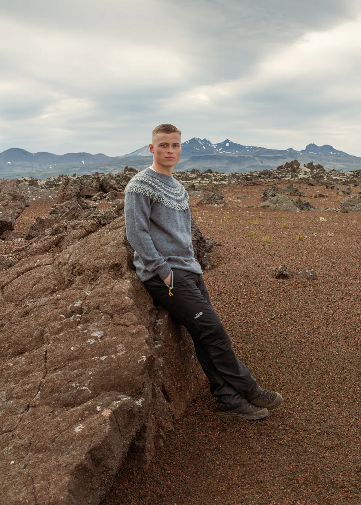 Un homme se tient près d’un rocher, une grande zone montagneuse derrière lui